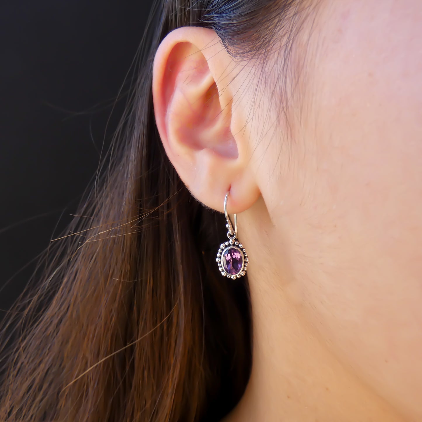 Woman wearing silver earrings with oval purple amethyst gemstones.
